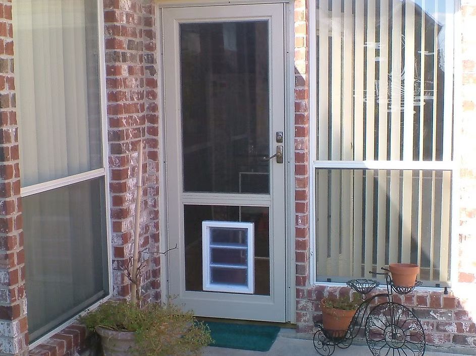 Exterior door with a pet door, surrounded by brick and windows with blinds.