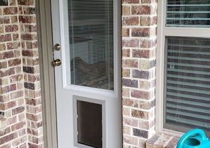 A white door with a dog door built into it is set in a brick wall. Window is adjacent.