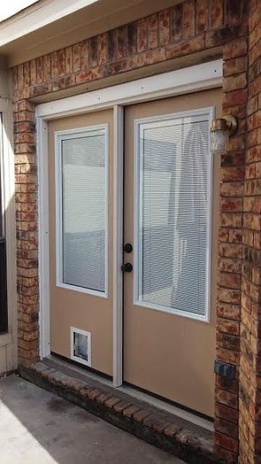 Tan double doors with white trim and a cat door, set in a brick exterior.
