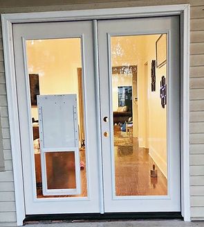 White double glass doors with a dog door on the left, reflecting a warm interior.
