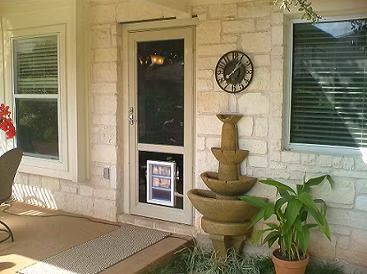 A cream-colored door with a doggy door, beside a tiered fountain and windows on a stone wall.