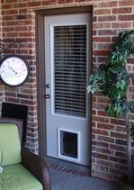White door with built-in pet door and window, set in a brick wall. A clock and chair are nearby.