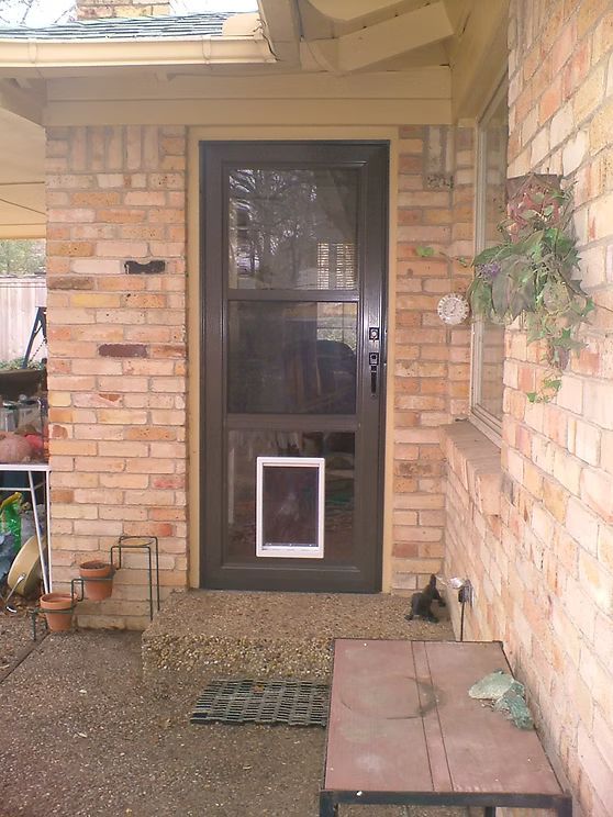 Brown screen door with doggy door, set in a brick entryway with a small bench and potted plants.