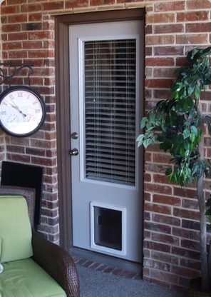 A white door with a dog door, a large window, and blinds on a brick wall, with a clock and chair.