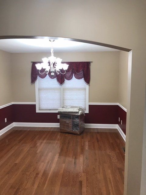 Empty dining room with wood floors, burgundy walls, chandelier, and a window with drapes.