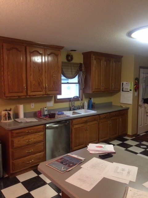 Kitchen with wooden cabinets, black-and-white tile floor, sink by window, and papers on a table.