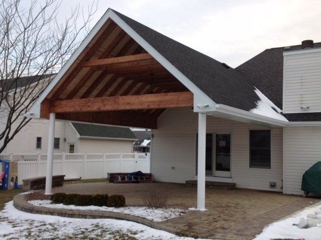Covered carport with wooden roof and white posts beside a beige house in a snowy yard