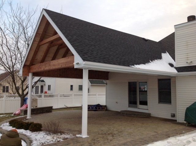 Covered patio attached to a beige house, with a peaked wood roof and white support posts in a snowy yard.
