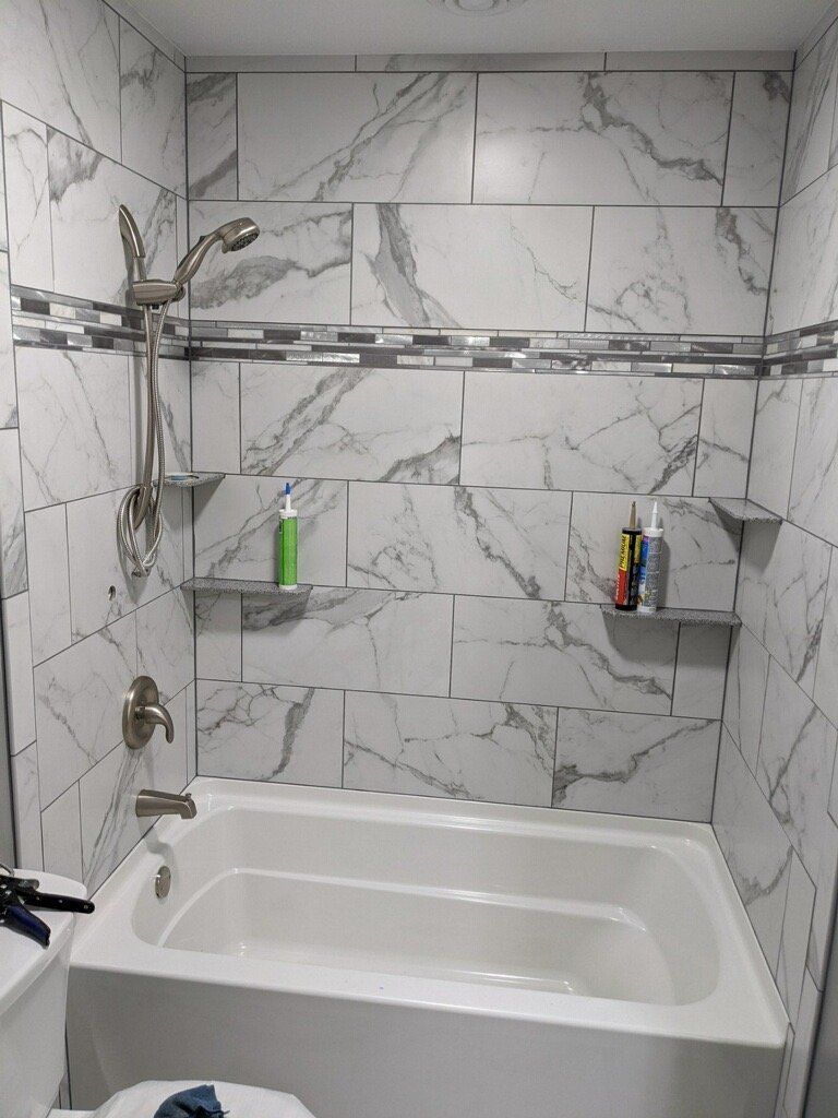 Empty tiled bathtub and shower with marble-pattern walls and wall-mounted shelf toiletries.