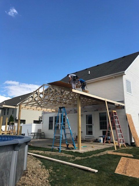Workers building a covered patio frame beside a white house, with ladders and lumber in the yard.