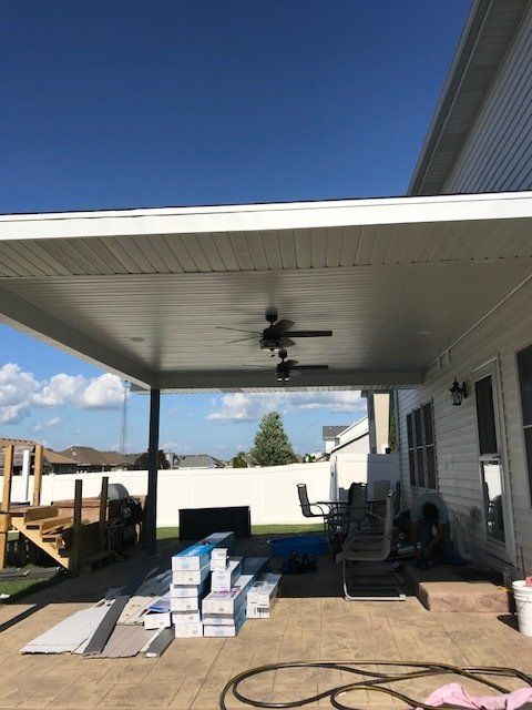 Covered patio under construction with stacked boxes, tools, and a ceiling fan overhead.
