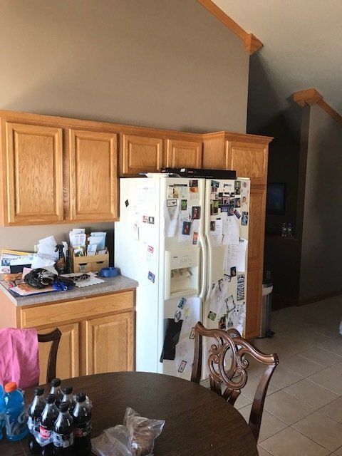 Kitchen with wooden cabinets, white fridge covered in magnets, and a dark hallway beside tiled flooring.