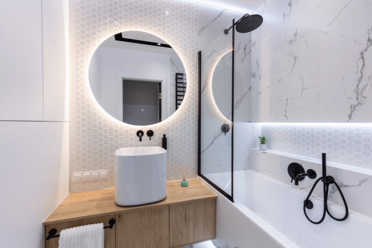 Modern bathroom with round lit mirror, wooden vanity, white sink, and black shower fixtures