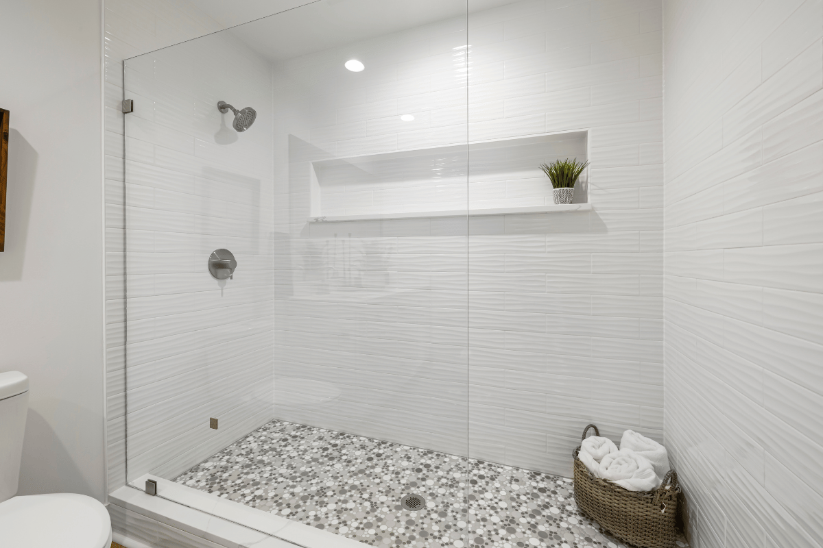 Modern white bathroom shower with glass door, pebble floor, shelf, and wicker basket