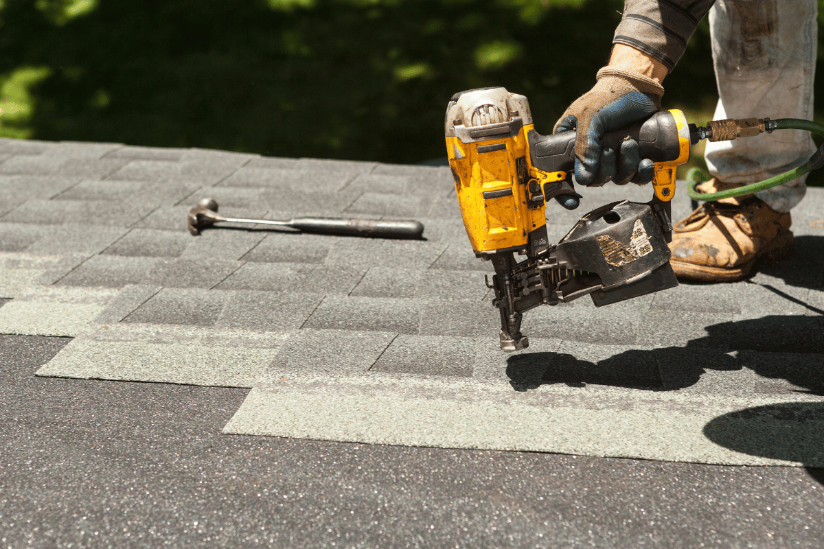 Worker using a yellow roofing nail gun to install shingles on a roof