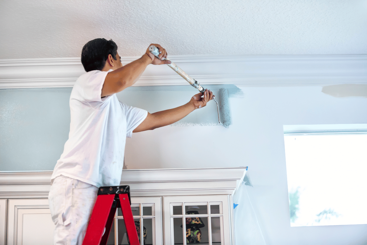 Person painting a ceiling with a roller on a red ladder in a bright room