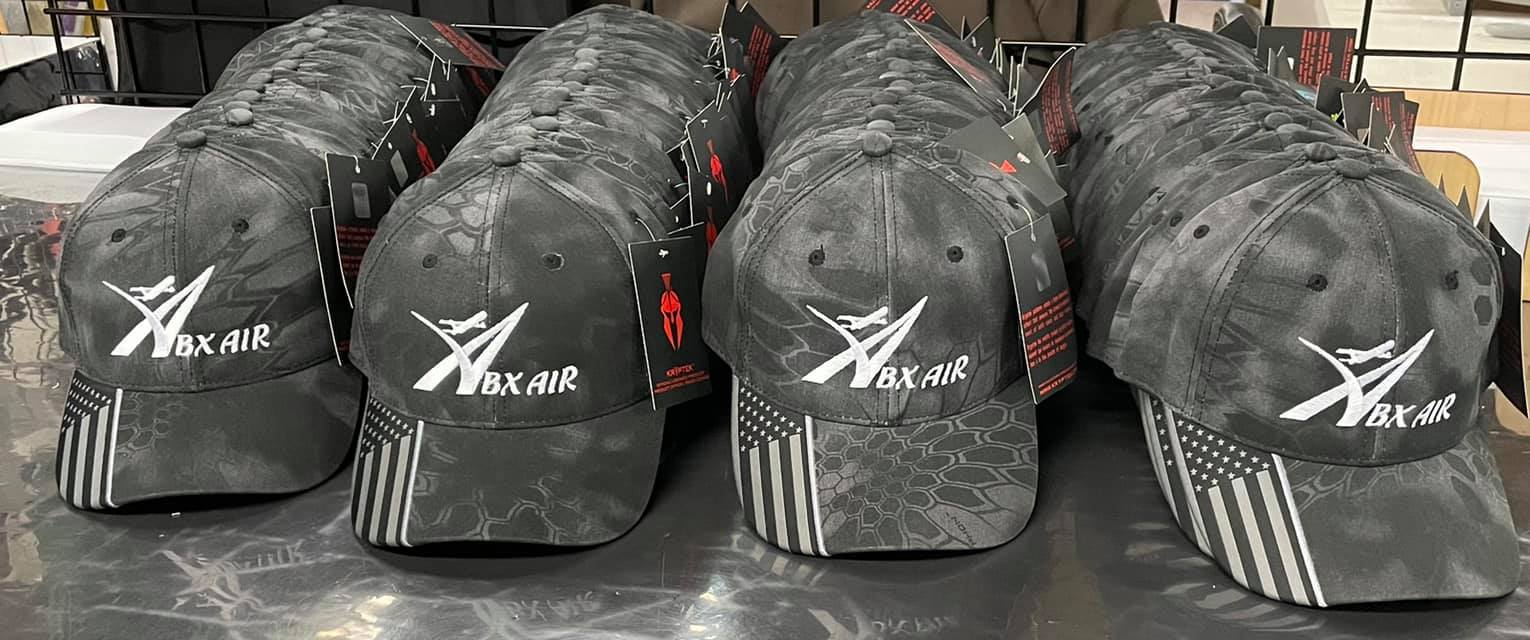 Four grey camouflage baseball caps in a row, each featuring a white logo and an American flag pattern on the brim.