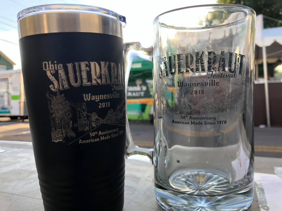 A black tumbler and a clear glass beer mug, both branded with the 2019 Ohio Sauerkraut Festival logo, sit on a table.