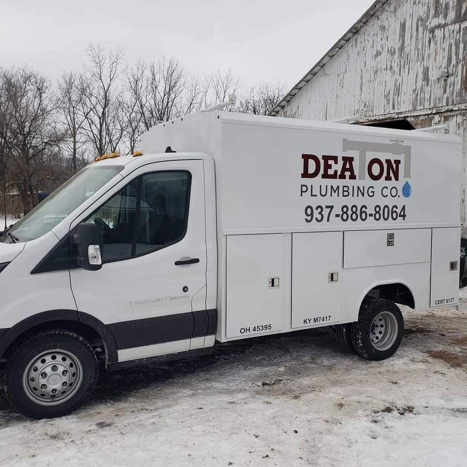 A white Deaton Plumbing Co. service truck parked on snow-covered ground in front of a white barn.