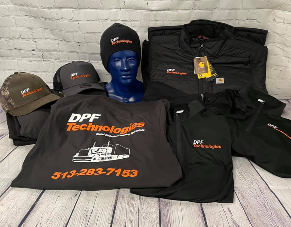 Black apparel and hats featuring the DPF Technologies logo and phone number, arranged on a white wood-grain surface.