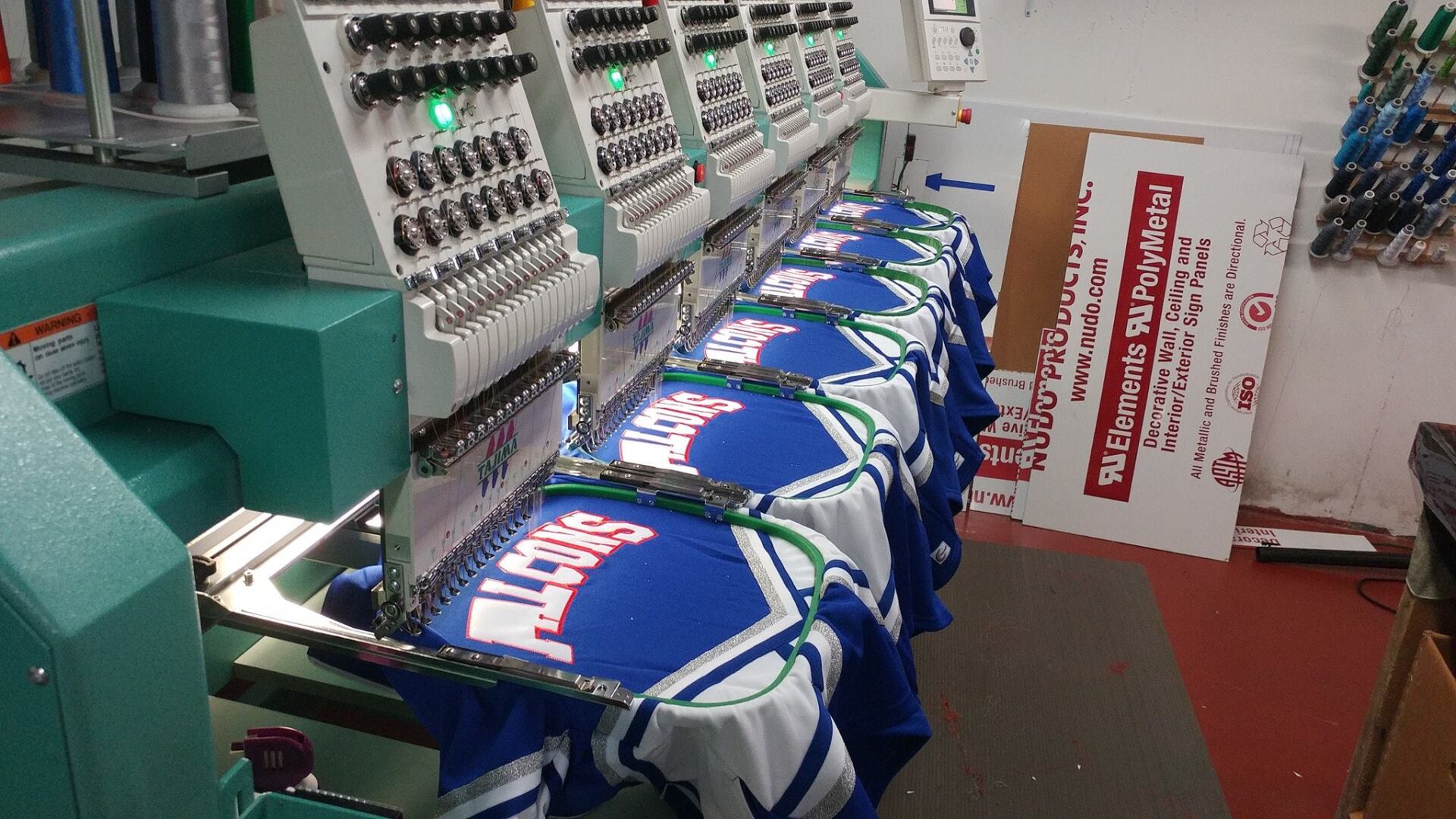 An industrial embroidery machine stitching blue and white jerseys in a production workshop.