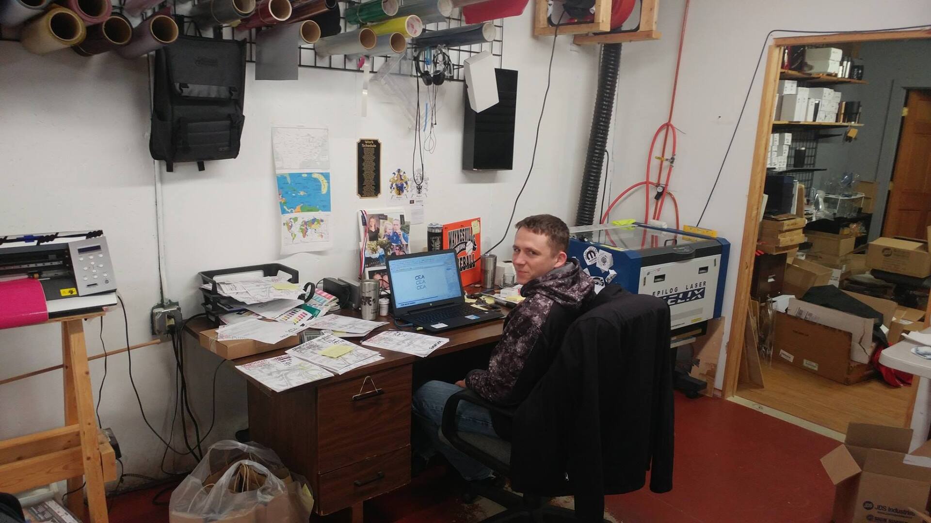 A person sitting at a cluttered desk with a laptop in a workspace filled with rolls of vinyl, tools, and a laser cutter.
