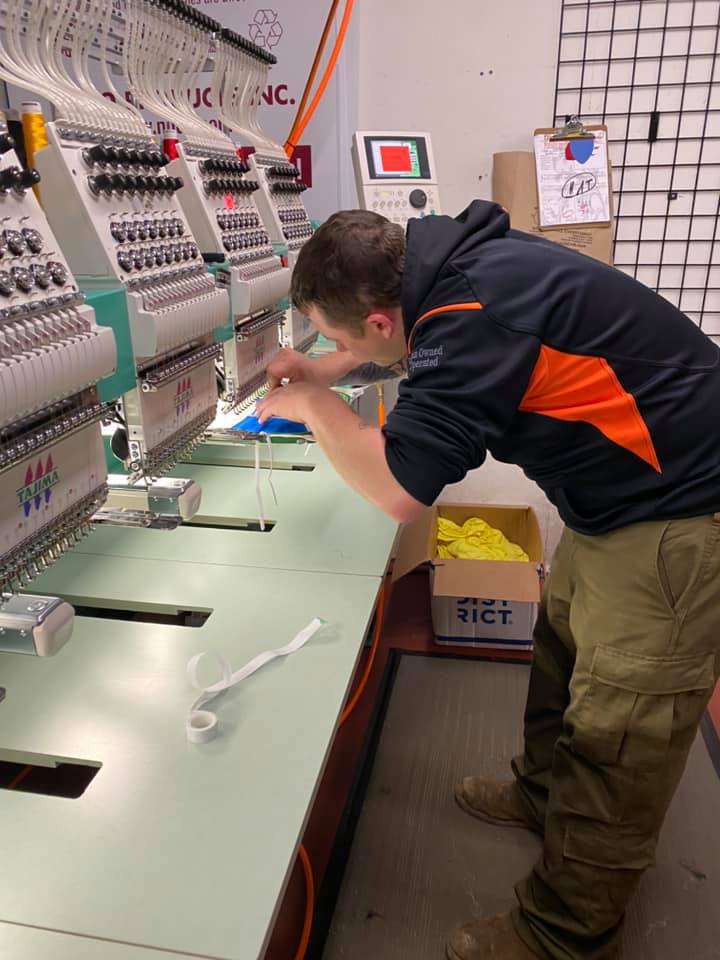 A person in a black-and-orange hoodie operates a large commercial embroidery machine in a factory setting.