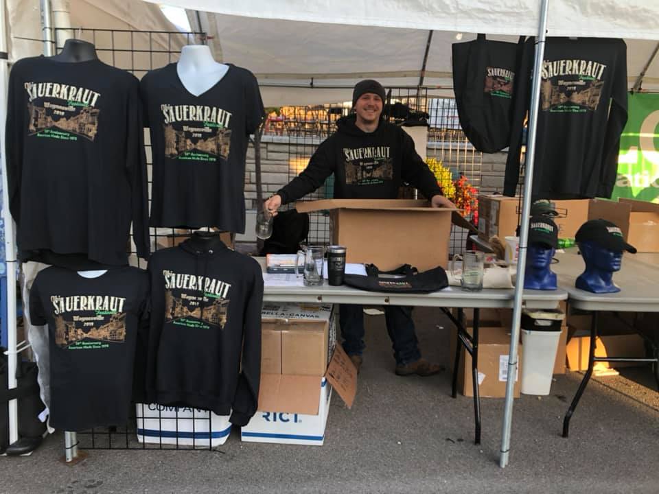 A person stands behind a table at an outdoor market stall selling black apparel and merchandise with a printed logo.