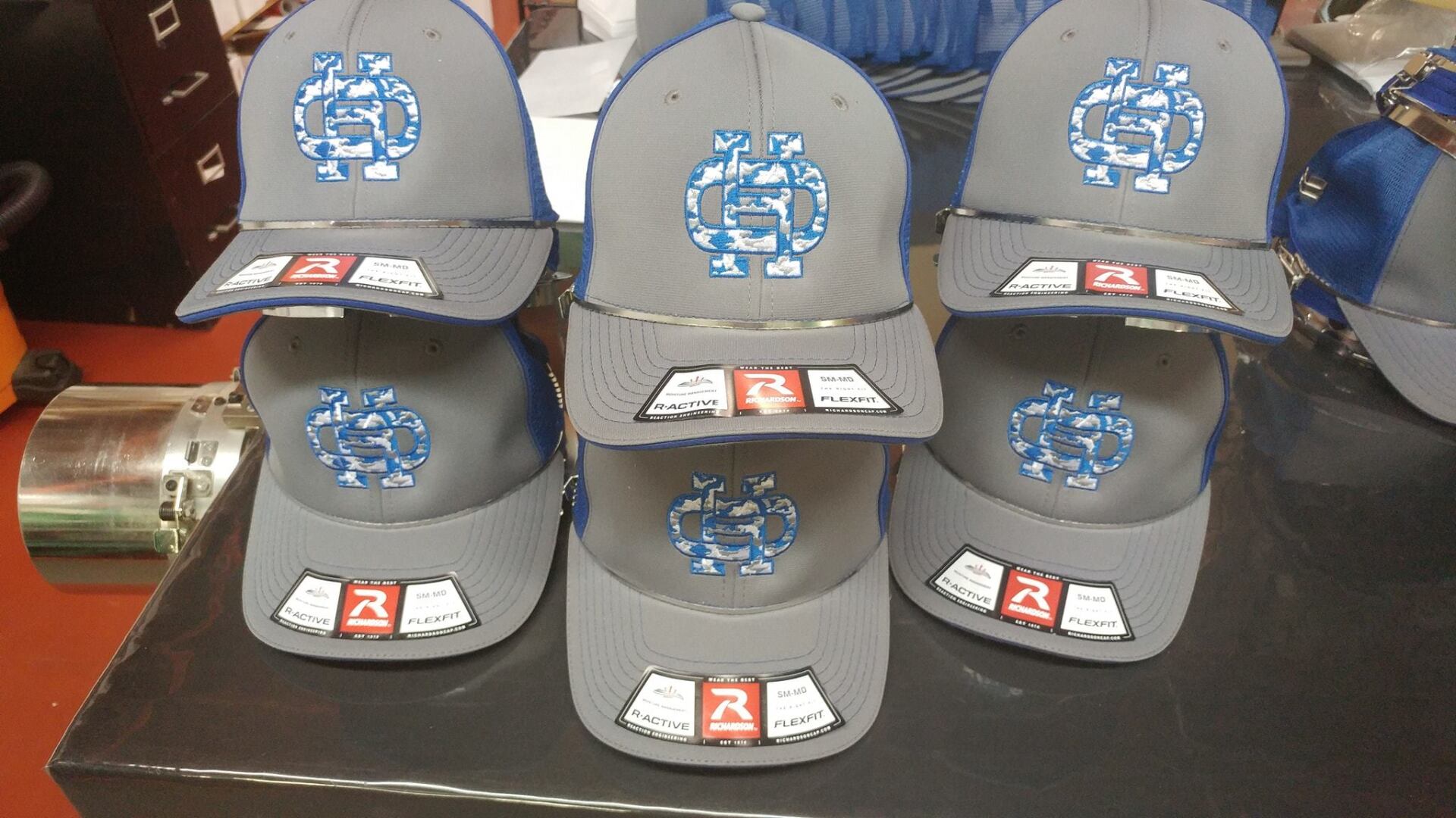 A stack of grey baseball caps featuring a blue camouflage logo and branding stickers, displayed on a black surface.