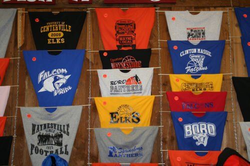 A display wall featuring many school spirit t-shirts in various colors and designs hung from wooden dowels.