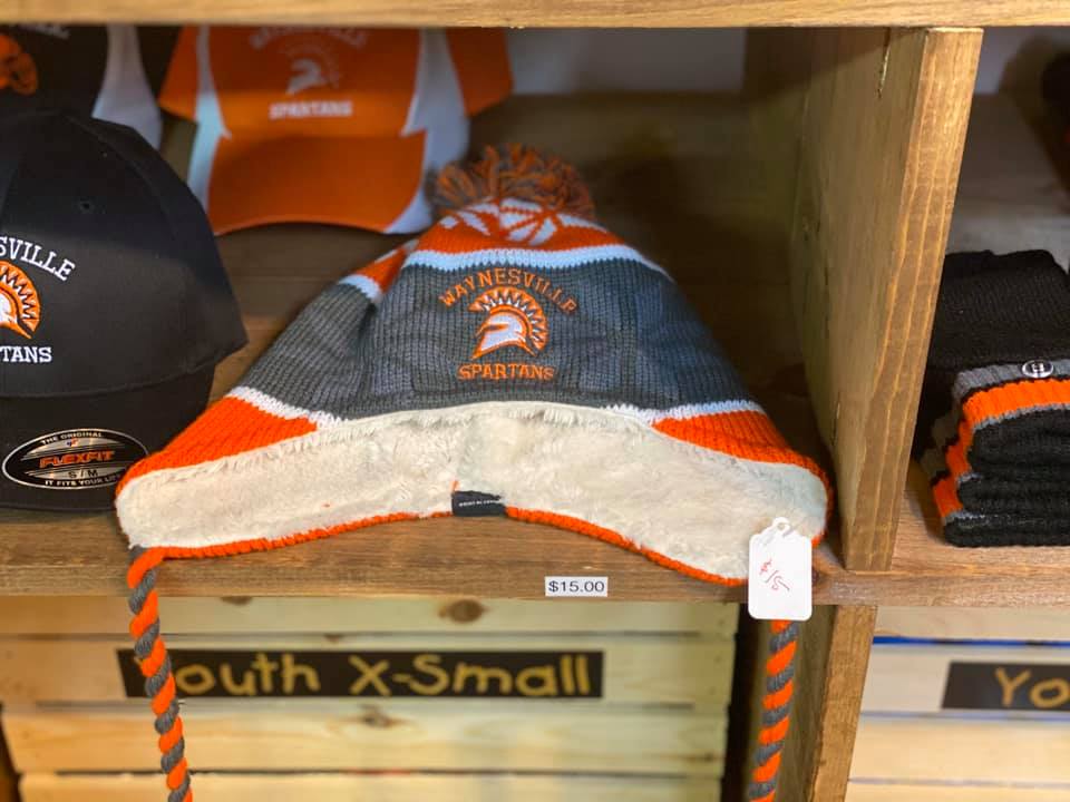 A grey, orange, and white Spartan beanie with earflaps and braids, displayed on a wooden shelf in a retail store.