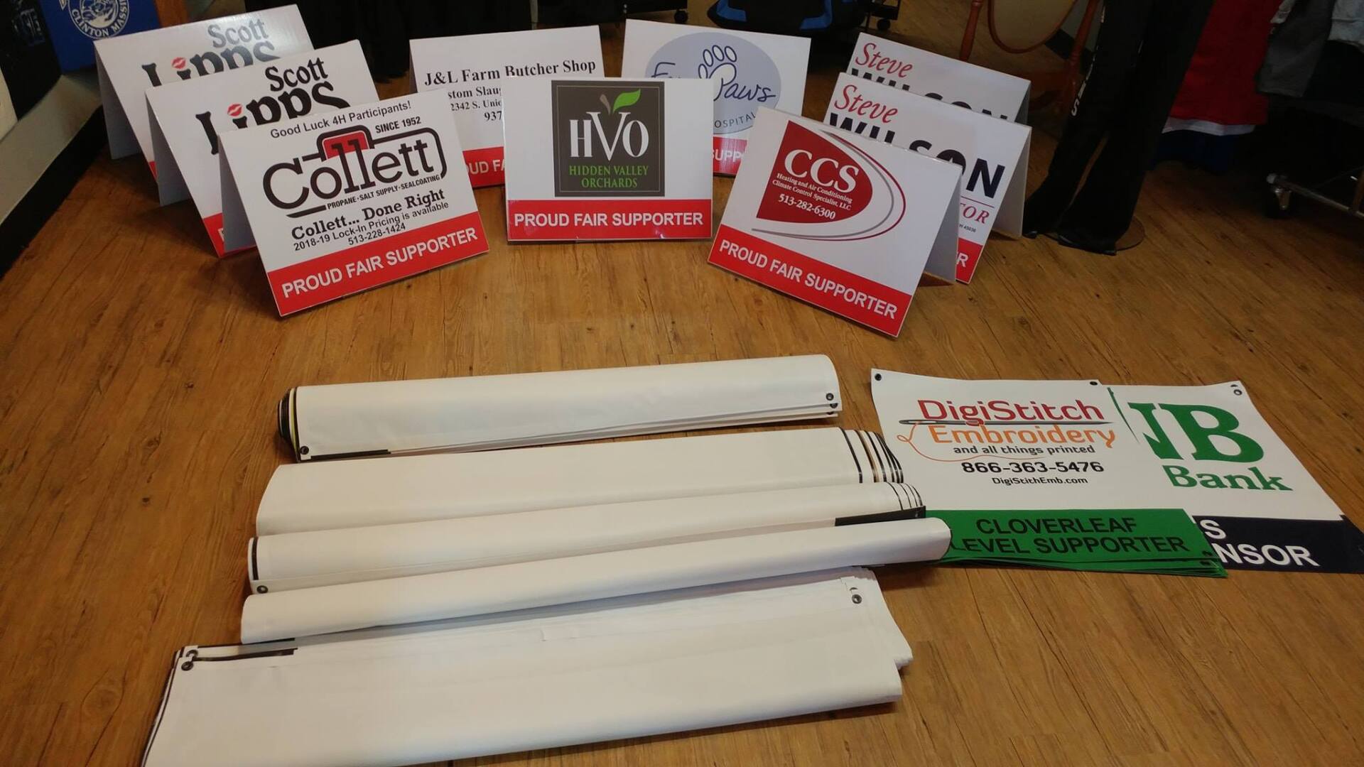 A collection of event sponsor signs and rolled-up banners displayed on a wooden floor.