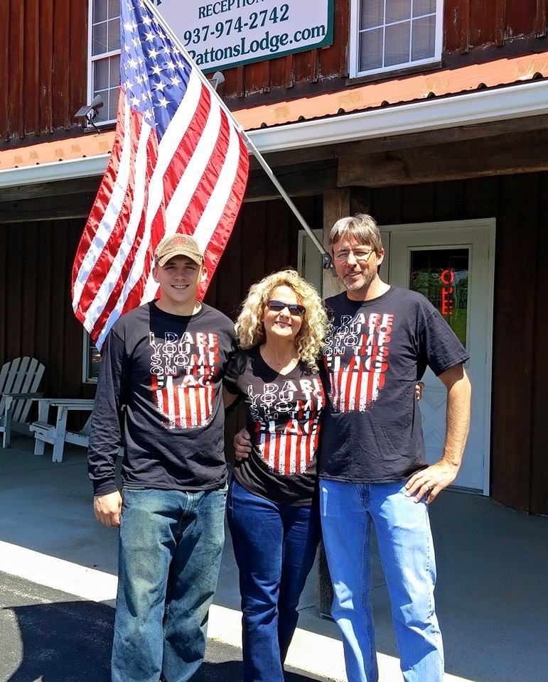 Three people wearing matching flag-themed shirts stand in front of a building with an American flag flying above them.
