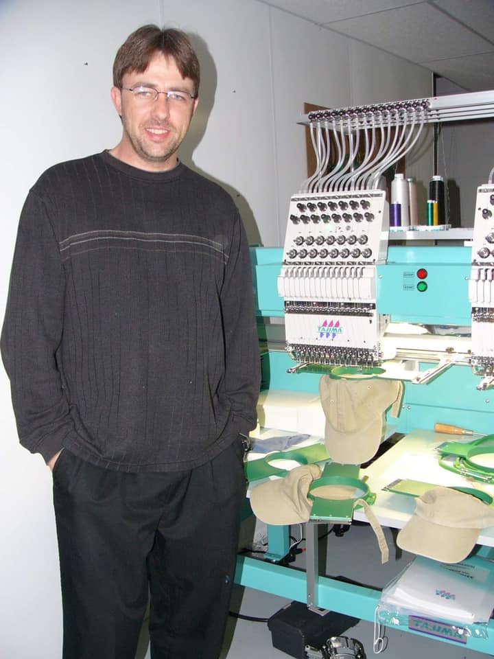 A person stands next to a multi-needle embroidery machine with tan caps in the hoops.