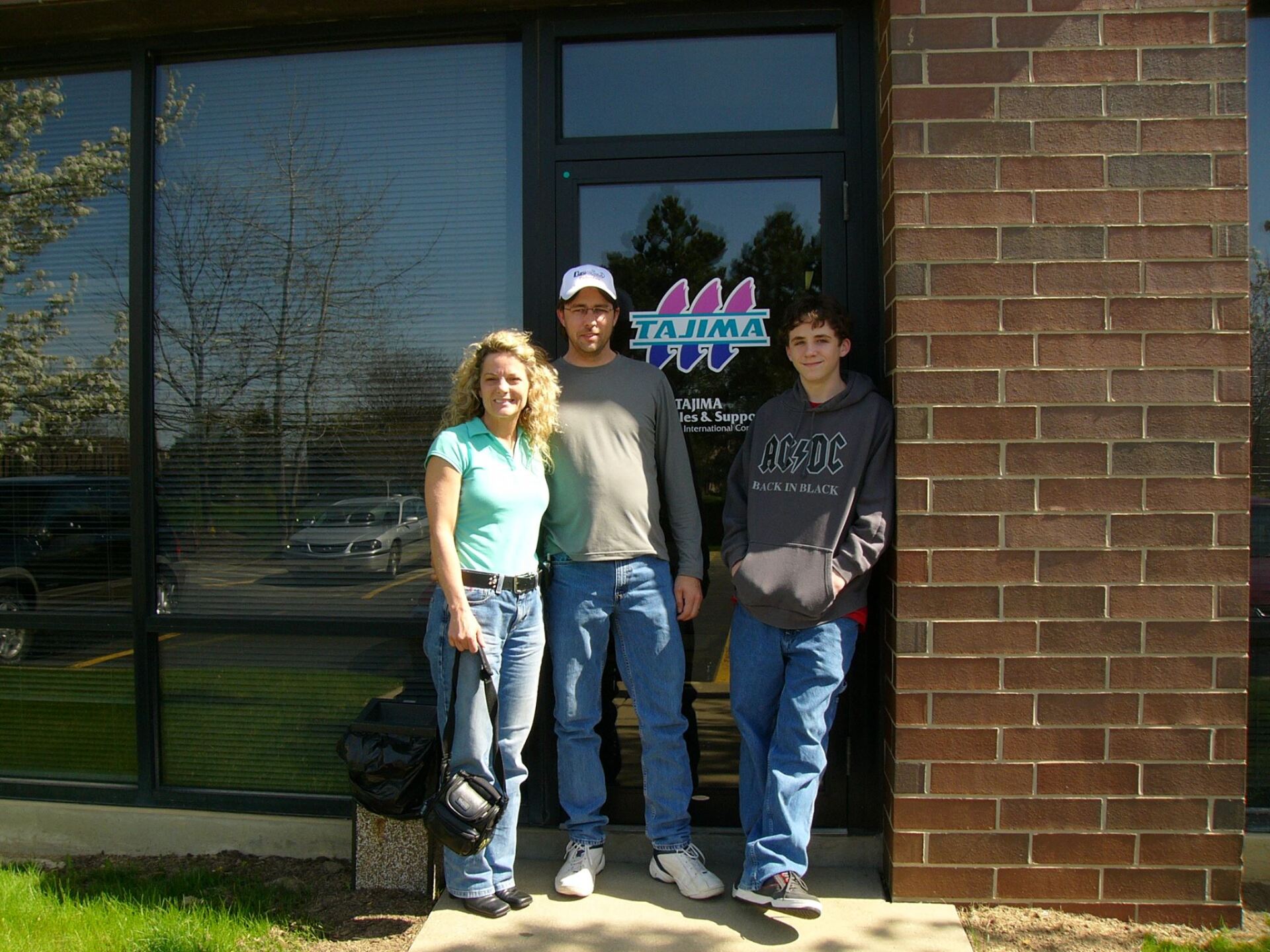 Three people stand smiling outside a brick building near a glass entrance door featuring a purple and blue sign.