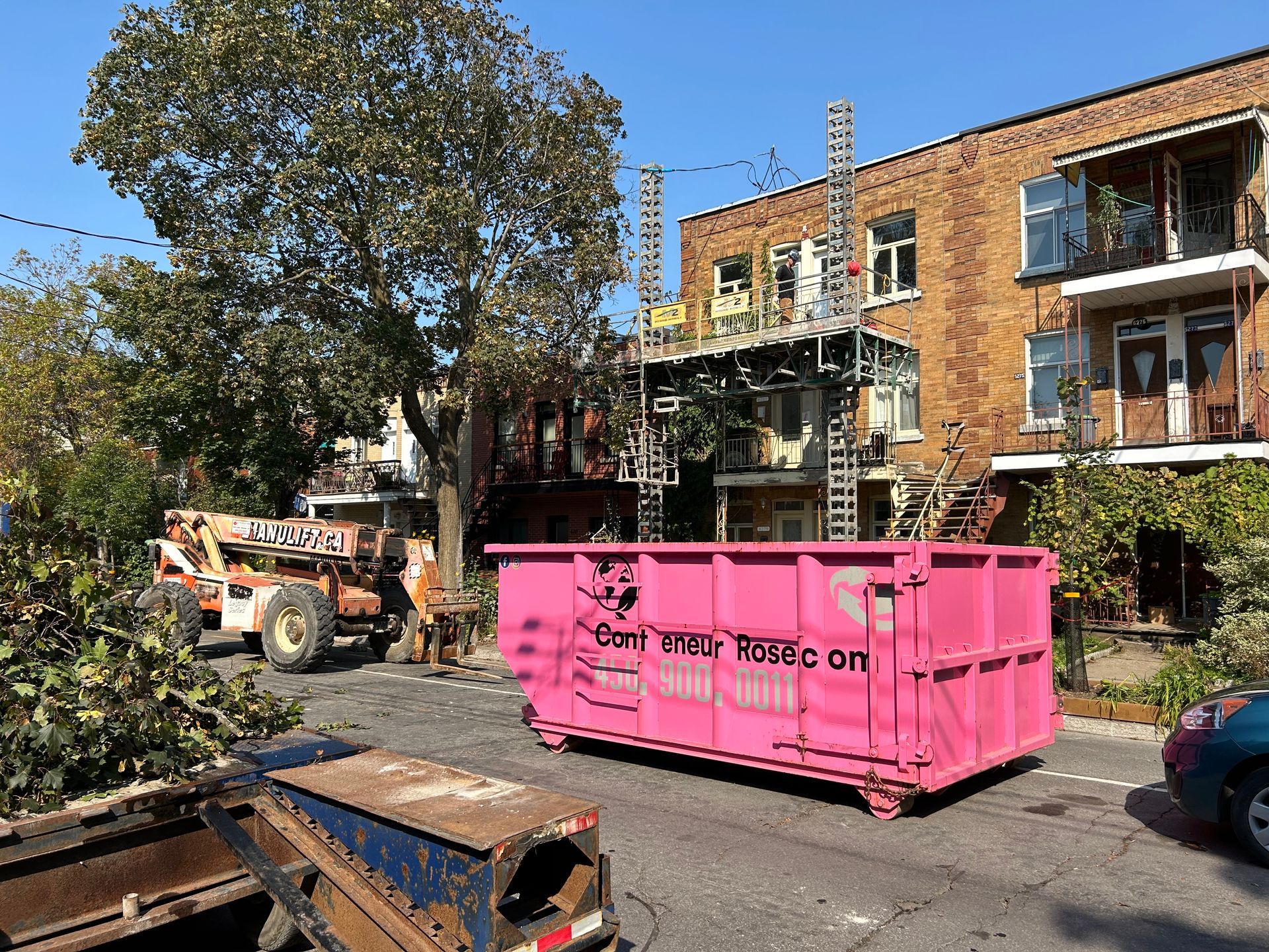 Chantier de construction avec une benne rose, des machines orange et un bâtiment en briques marron.