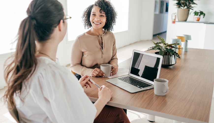 Duas mulheres estão sentadas à mesa com um laptop e tomando café.