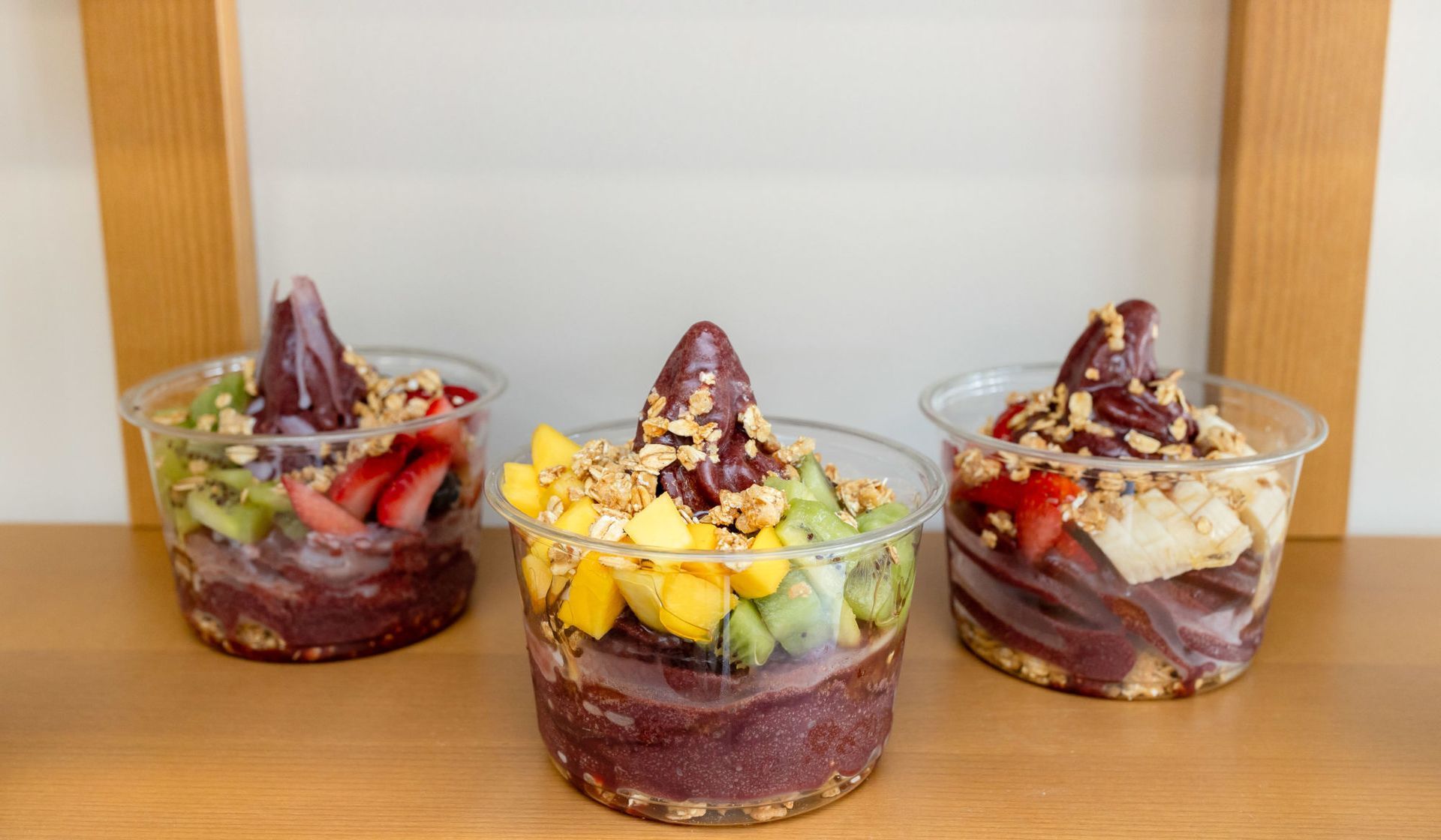 Three cups of açai bowls are sitting on a wooden table.