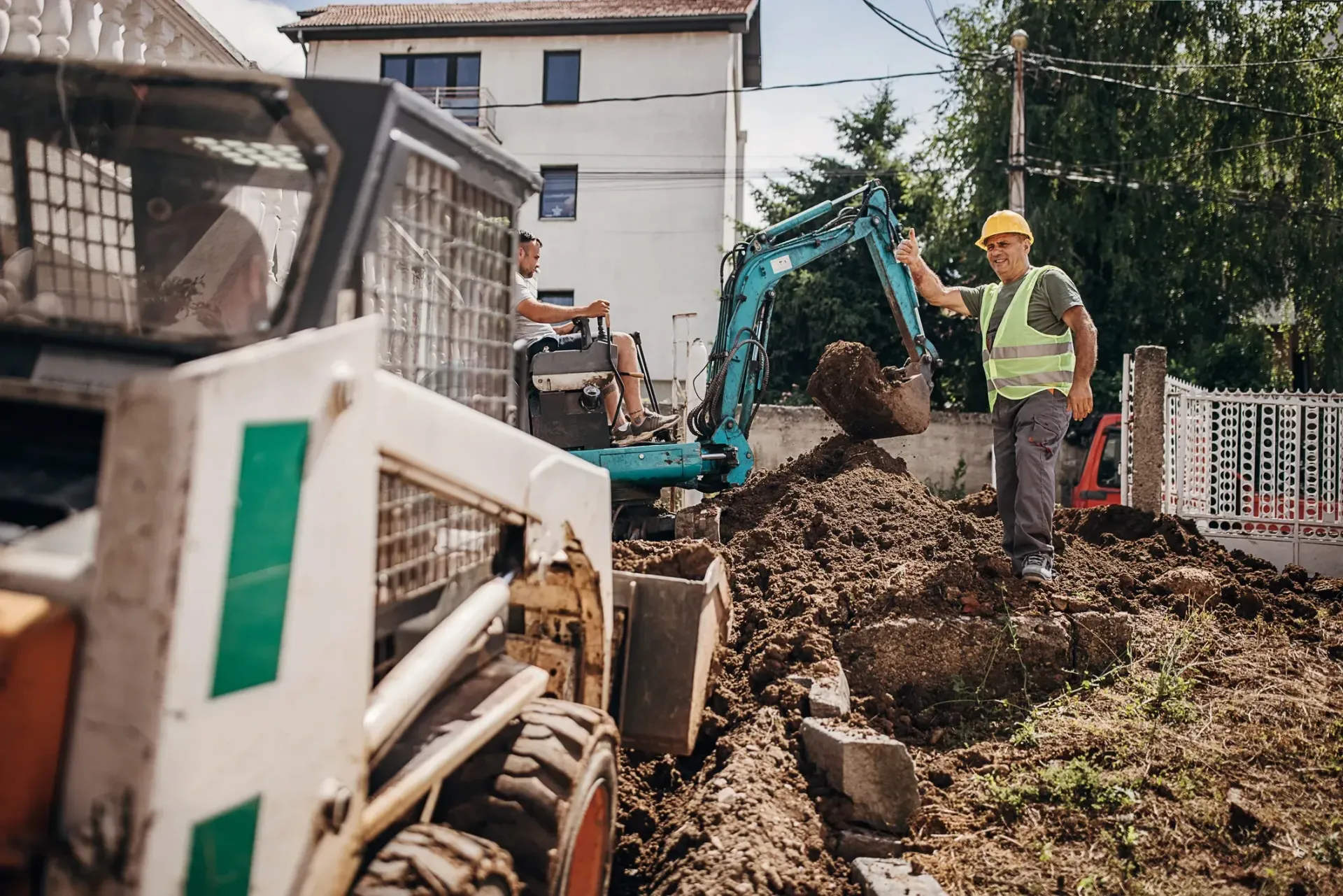 Construction worker by excavator clearing muddy debris beside a building