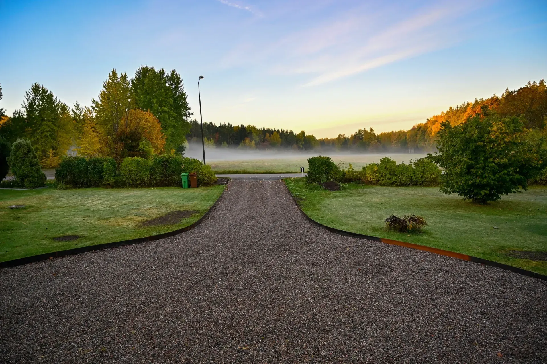 Gravel driveway leading through a misty garden at sunrise, with trees and colorful shrubs on both sides