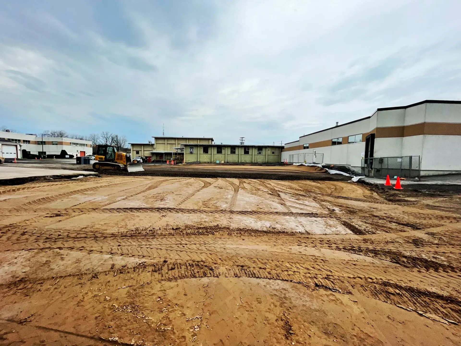 Large muddy construction site beside warehouses under an overcast sky
