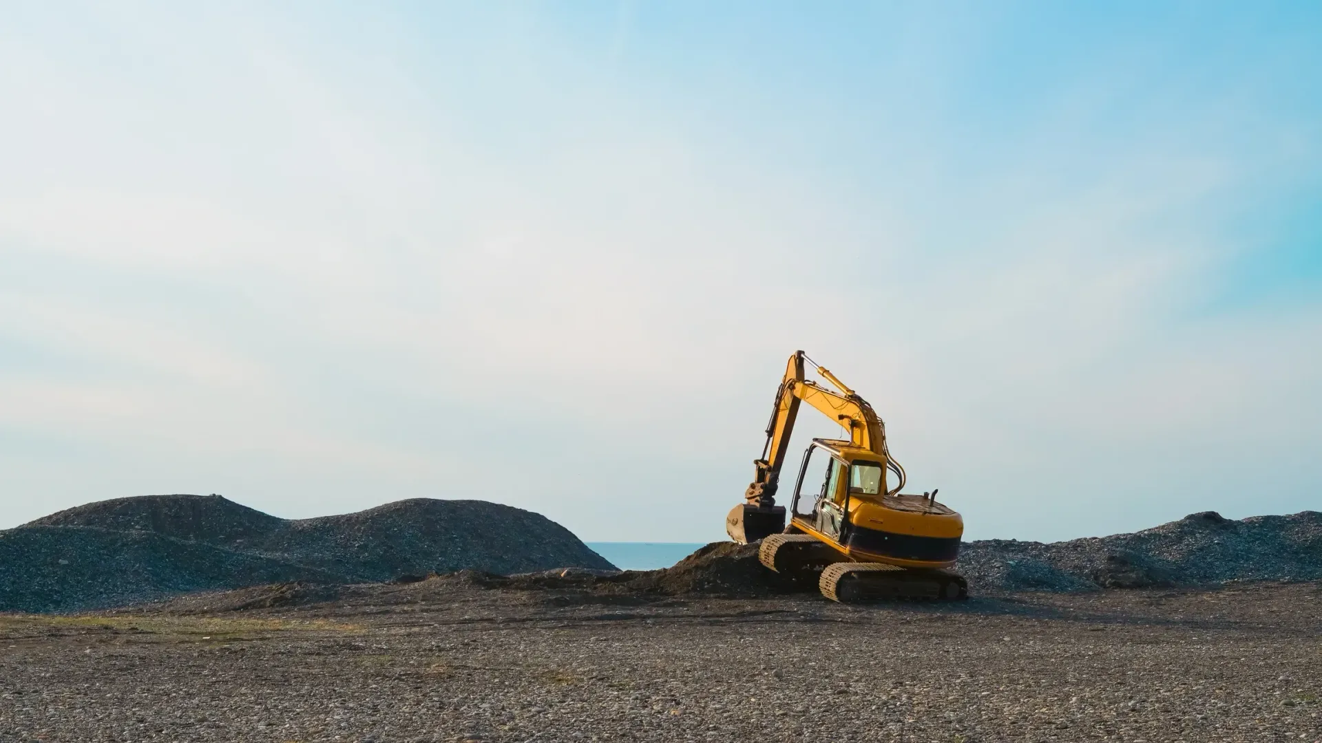 Yellow excavator on a gravel lot with dirt mounds under a blue sky