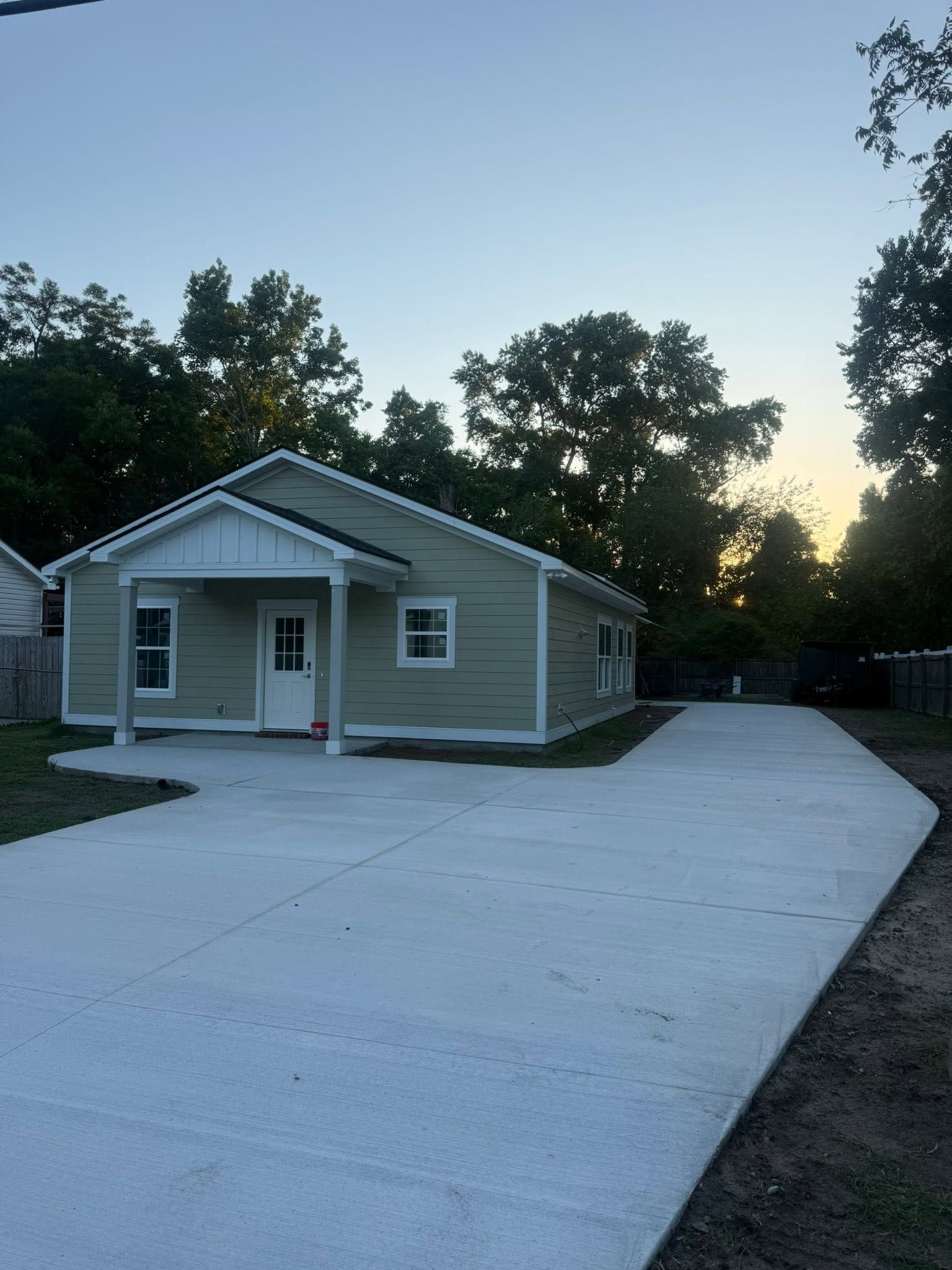 Small gray house with a covered porch and a long concrete driveway at dusk.