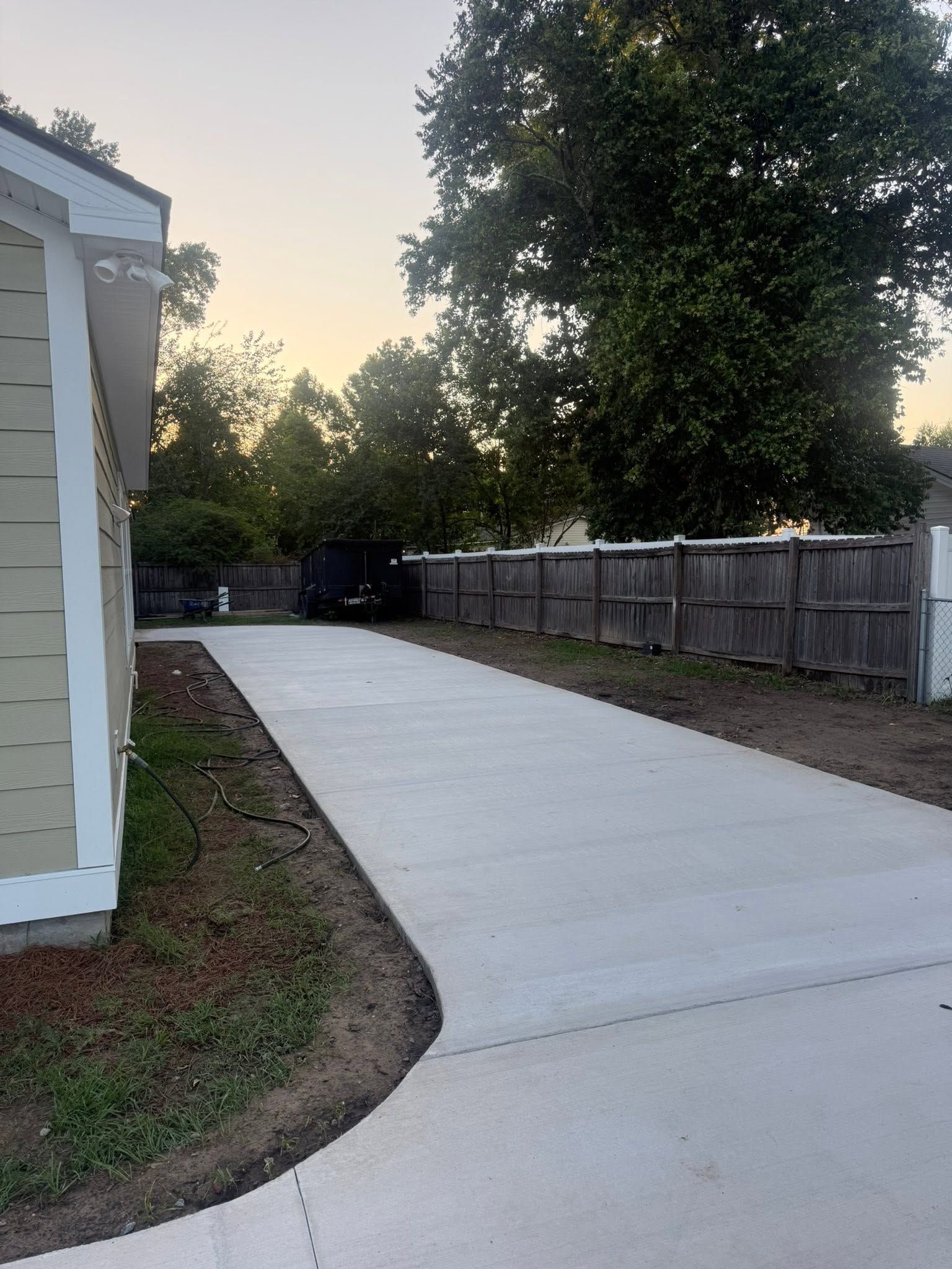 Concrete driveway curving past a house, with a wooden fence and trees at dusk.