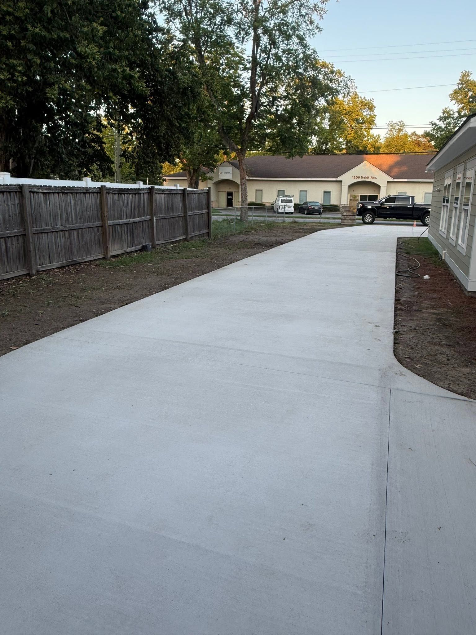 Freshly poured concrete driveway leading to a beige house, with a wooden fence and parked vehicle in the background