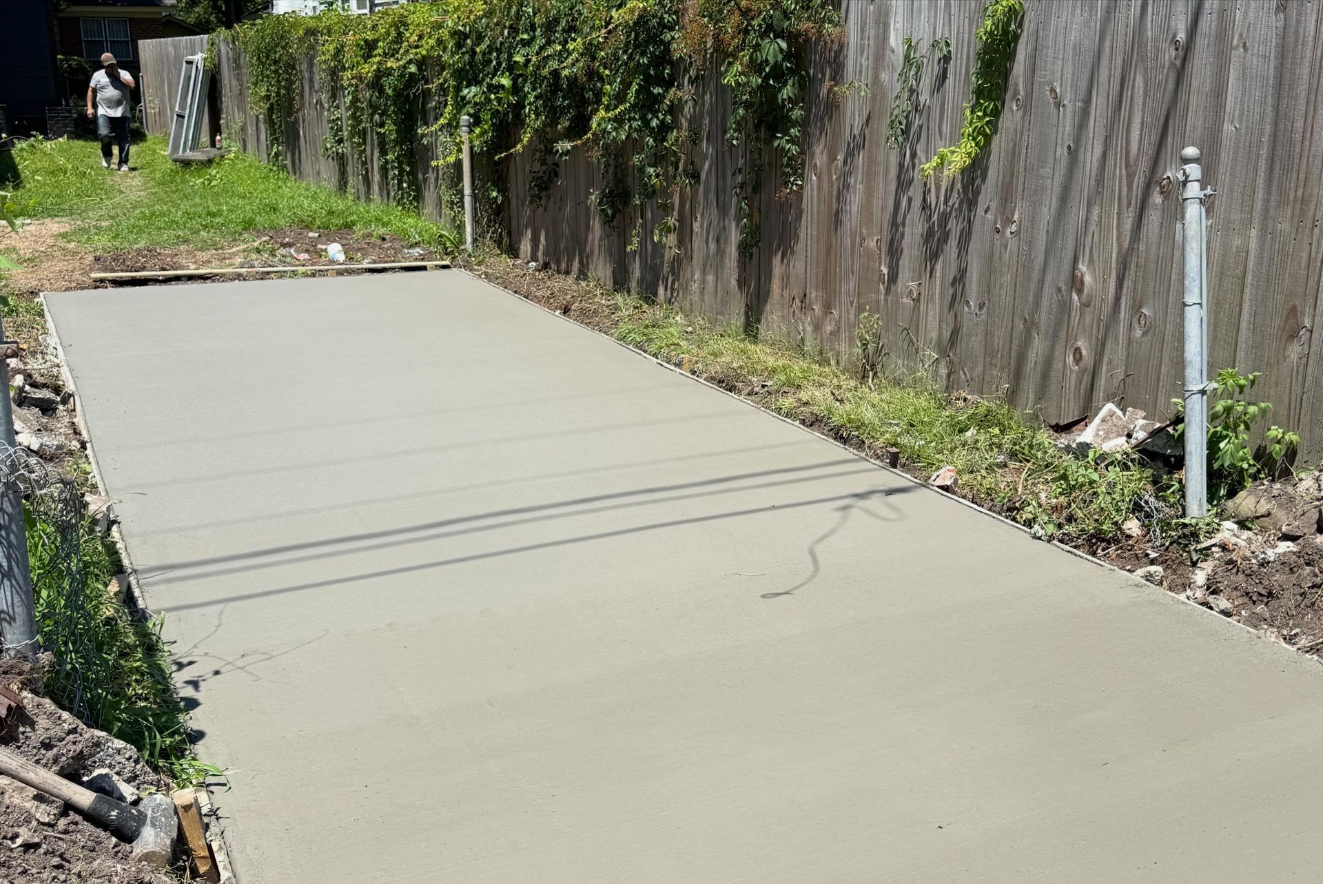 Freshly poured concrete driveway beside a wooden fence and greenery.