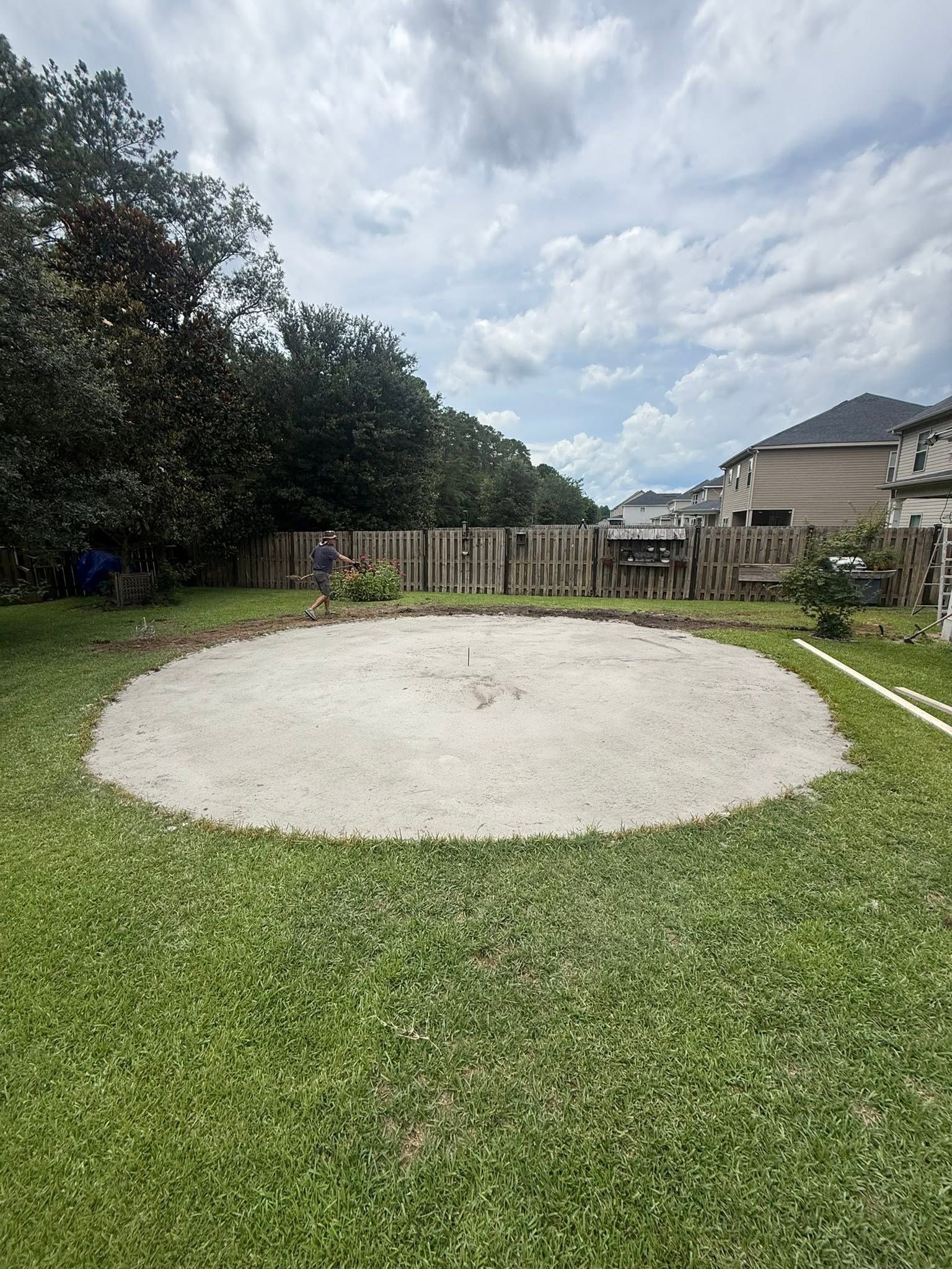 Backyard with a circular sand play area, green lawn, trees, and a fence under a cloudy sky