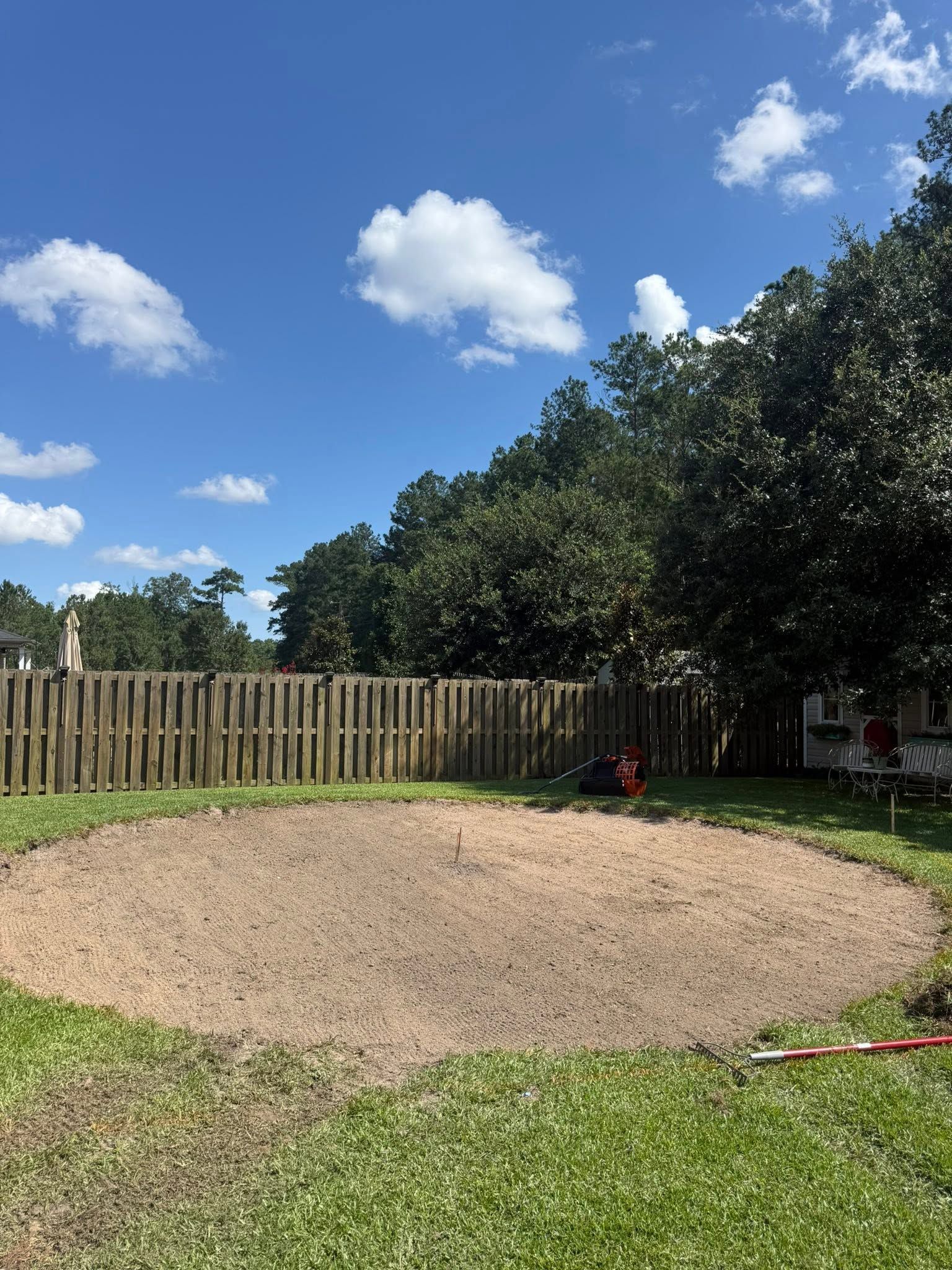Backyard with a large round gravel area, green lawn, wooden fence, and blue sky with clouds