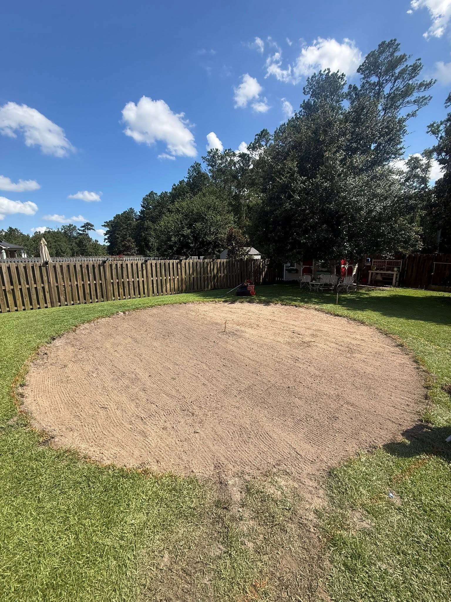 Circular gravel patio in a grassy backyard under a blue sky, with a wooden fence and trees in the background