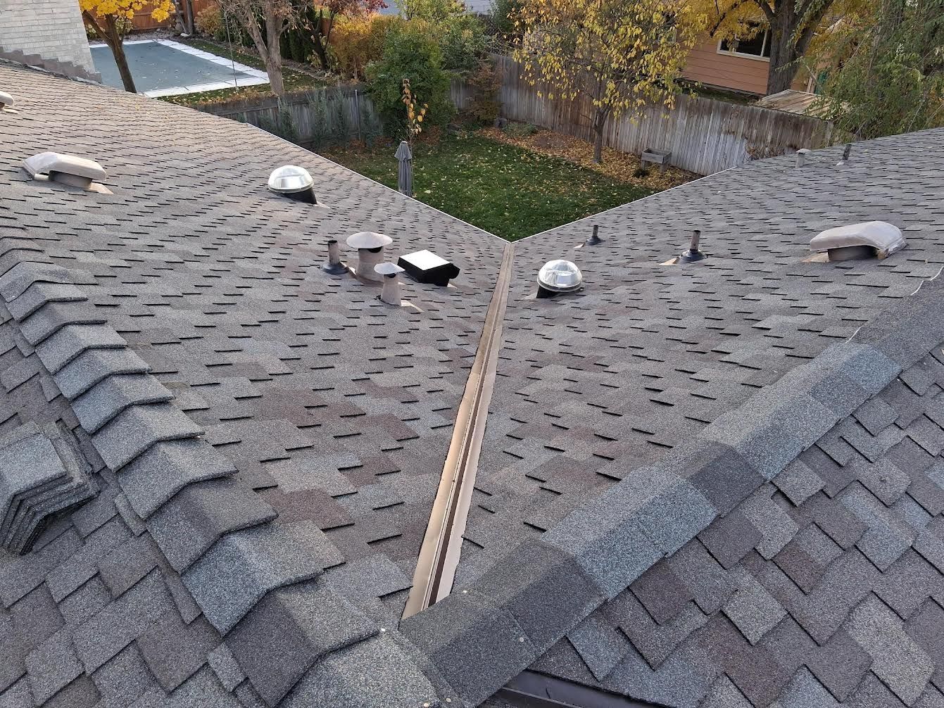 An elevated view of a gray asphalt shingle roof featuring a central valley, several roof vents, and a cap ridge.